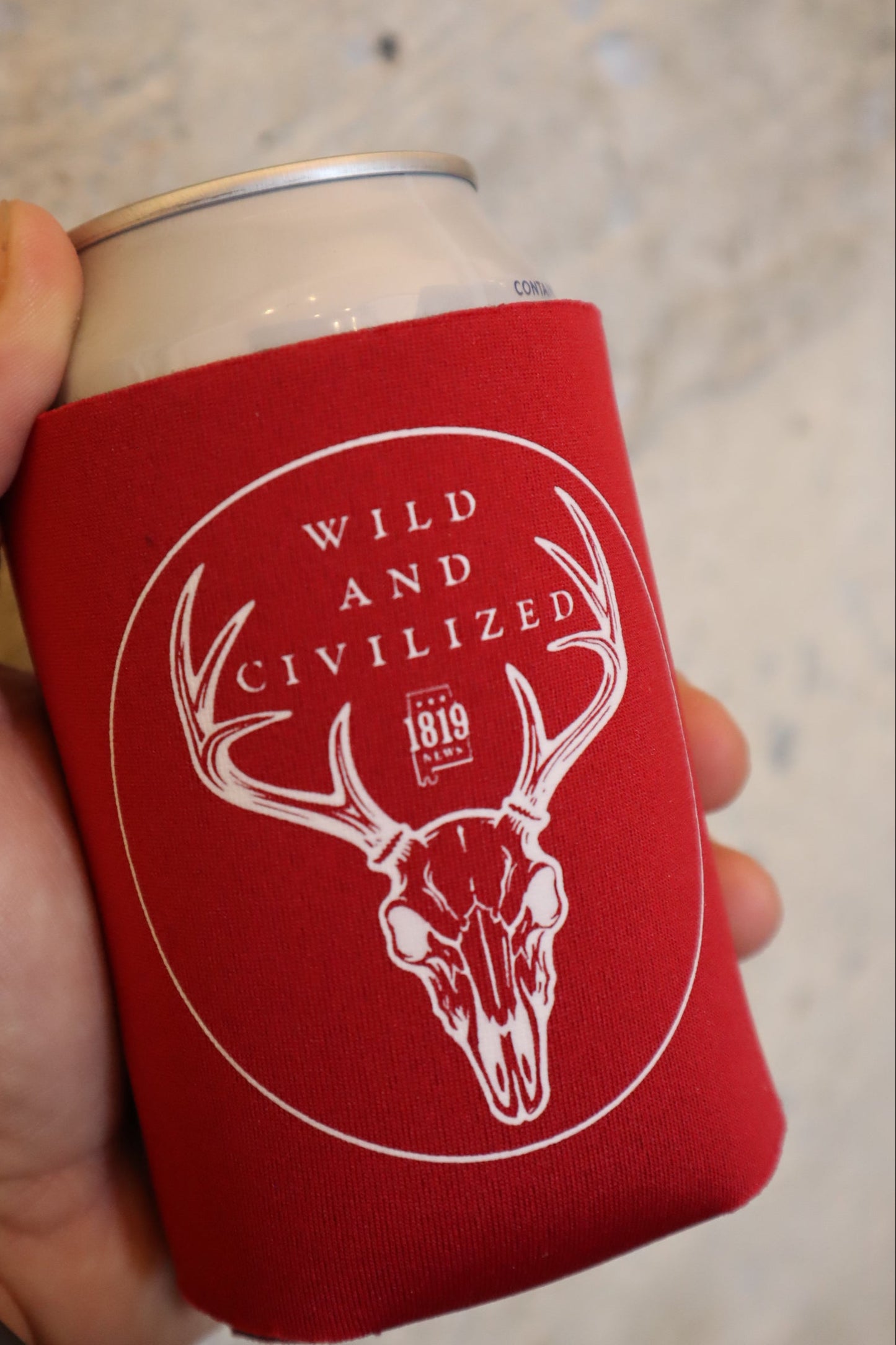 Red can cooler with white graphic and text held by a hand on a blurred background